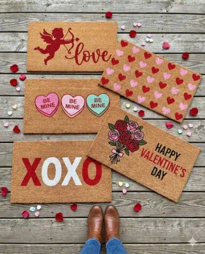 DIY Valentine’s Day doormats with heart patterns, Cupid design, XOXO text, Be Mine messages, and Happy Valentine’s Day lettering on coir mats displayed on a wooden porch.