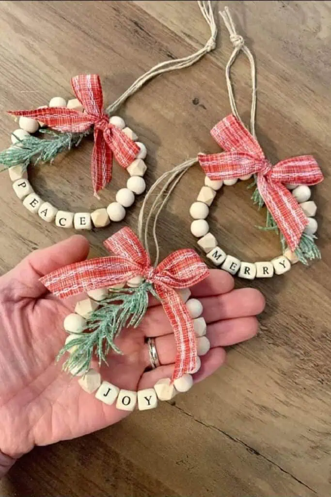DIY wood bead Christmas wreath ornaments decorated with red plaid bows, greenery, and letter beads spelling Peace, Joy, and Merry, perfect for rustic farmhouse holiday decor.
