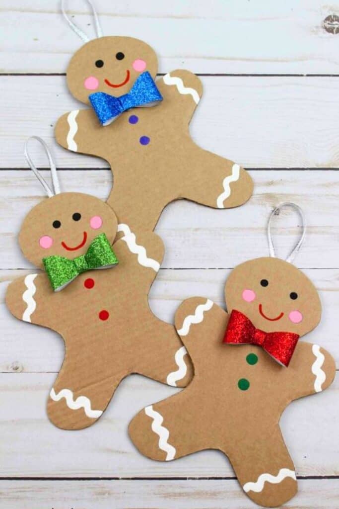 Three handmade gingerbread man Christmas ornaments made from cardboard, decorated with colorful glitter bows, pink cheeks, and white icing details, laid out on a white wood background. A fun and easy DIY holiday craft for kids or adults, perfect for a Candyland Christmas tree or classroom activity.