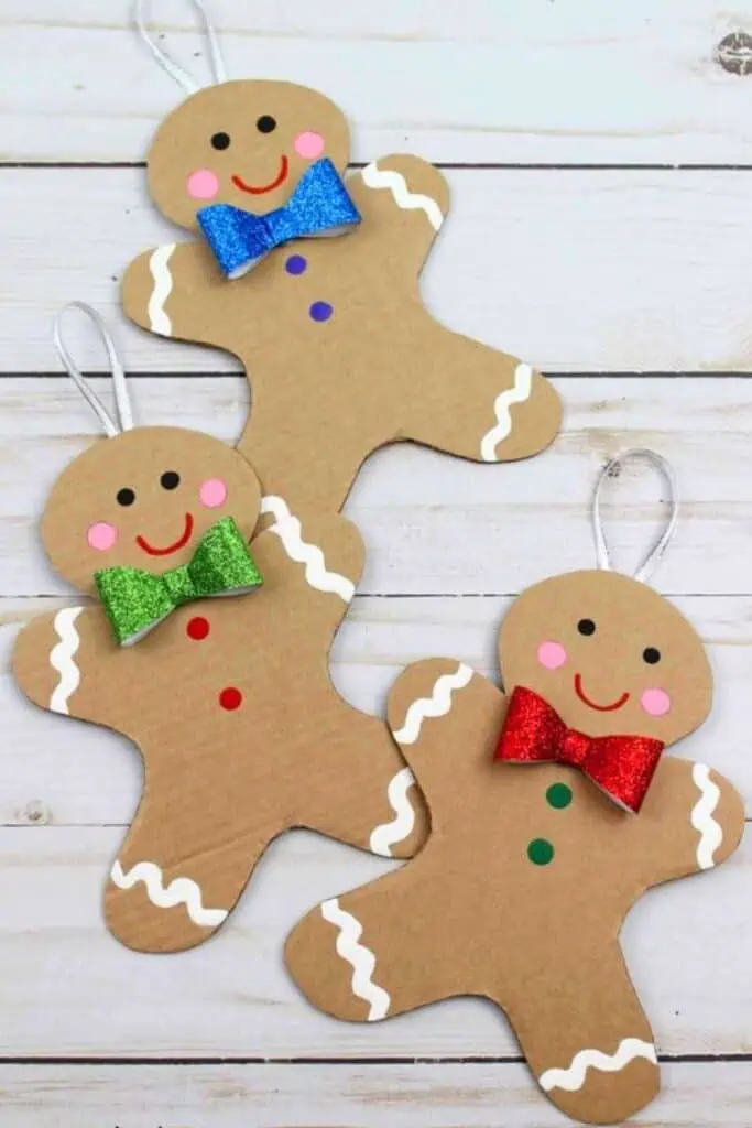 Three handmade gingerbread man Christmas ornaments made from cardboard, decorated with colorful glitter bows, pink cheeks, and white icing details, laid out on a white wood background. A fun and easy DIY holiday craft for kids or adults, perfect for a Candyland Christmas tree or classroom activity.