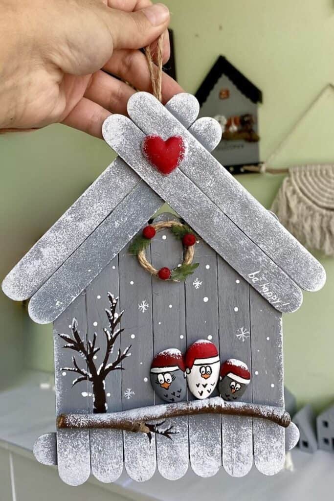 DIY popsicle‑stick Christmas house ornament with snowy roof, red heart, tiny wreath, and painted pebble birds in Santa hats, hung with jute twine.”
