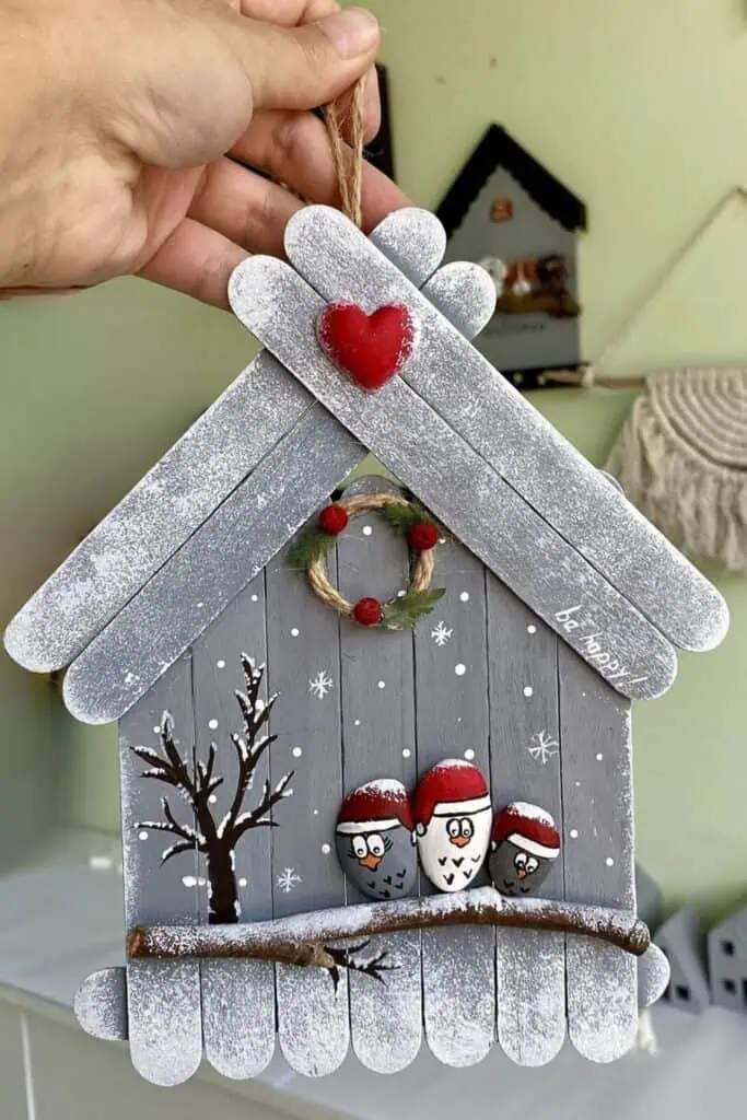 DIY popsicle‑stick Christmas house ornament with snowy roof, red heart, tiny wreath, and painted pebble birds in Santa hats, hung with jute twine.”