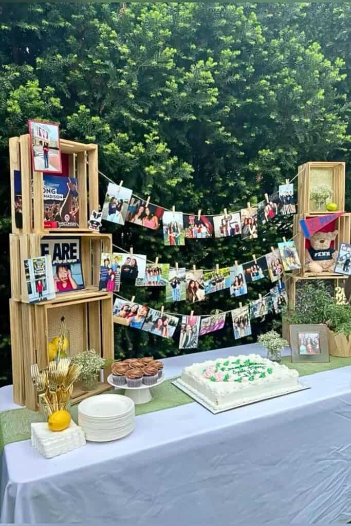 Outdoor graduation party college table with wood crates, a DIY photo banner display using clothespins, framed pictures, cupcakes, floral accents, and a decorated sheet cake—perfect for a backyard celebration.
