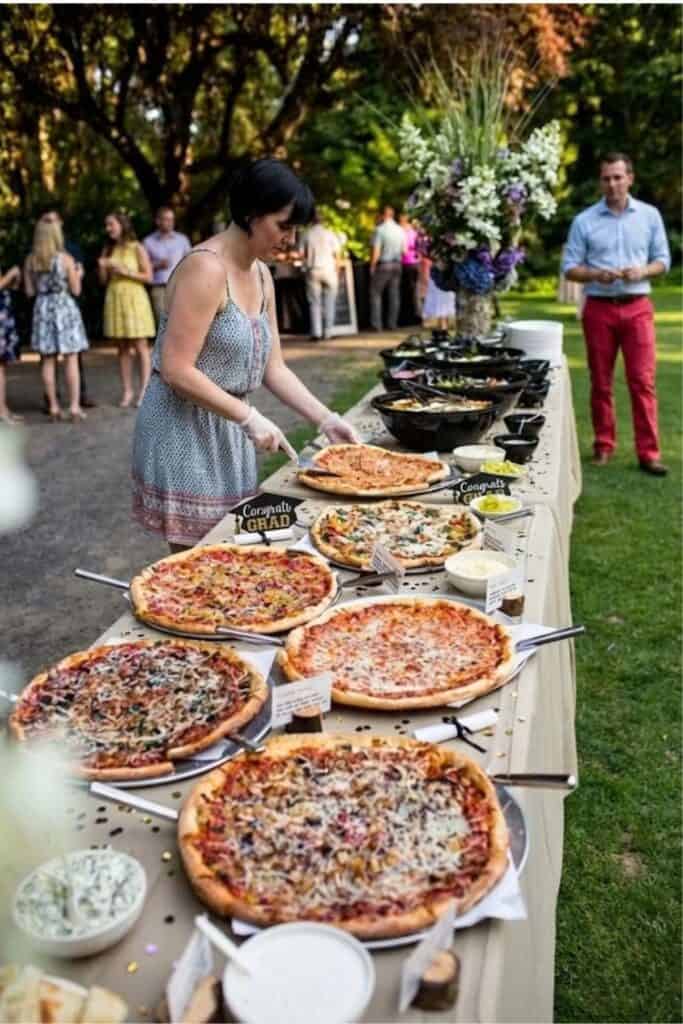 Person serving pizza at a graduation party buffet with various pizzas, salads, and a 'Congrats Grad' sign on the table