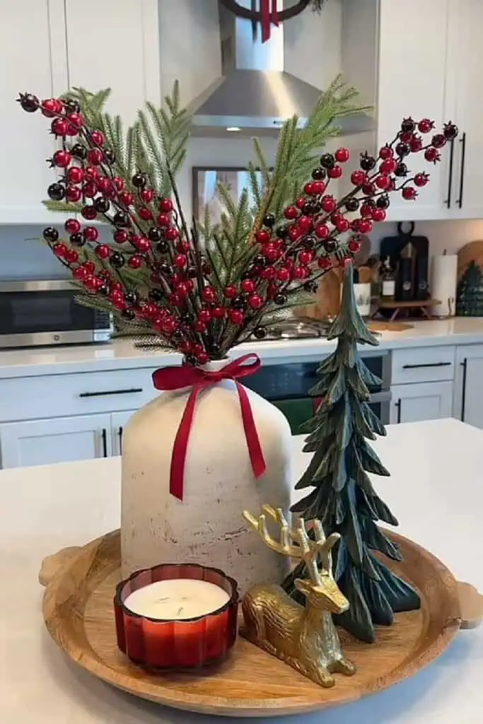 Farmhouse Christmas tray centerpiece on a kitchen island: round wood tray with a stoneware jug of red berries and pine tied with a red ribbon, green tree figurine, gold reindeer, and a red candle.