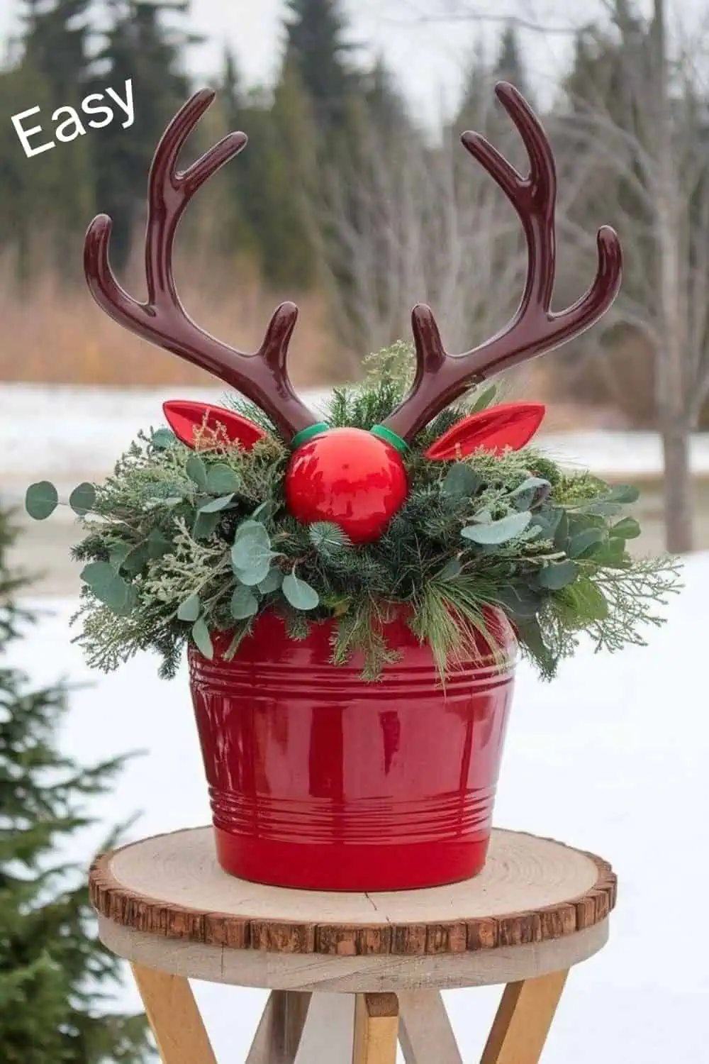 Red metal bucket styled as a reindeer with antlers, greenery, and a red ornament nose, displayed outdoors on a wood stand. A festive and rustic farmhouse glam Christmas decoration for porches or front doors.