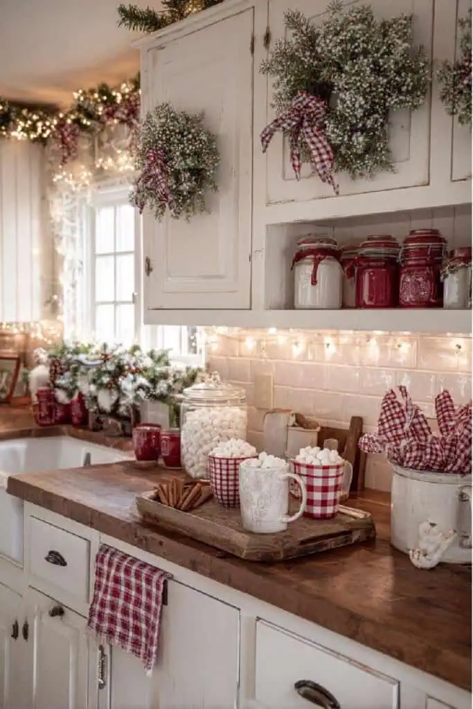 A cozy farmhouse kitchen decorated for Christmas with garlands, wreaths, red and white gingham accents, jars of marshmallows and cocoa, and warm twinkle lights. The rustic wooden countertops and vintage cabinets create a charming country Christmas atmosphere.