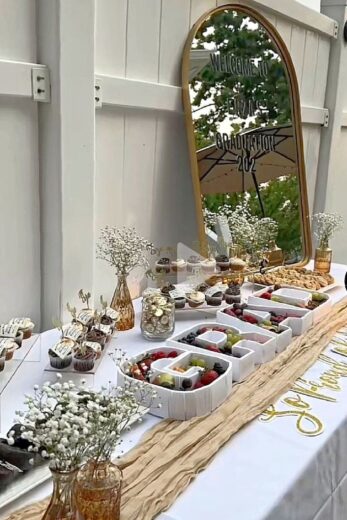 Elegant backyard graduation party dessert table with charcuterie cups, cupcakes, fresh fruit, gold accents, and a mirror welcome sign for an outdoor graduation celebration.