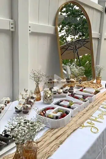 Elegant backyard graduation party dessert table with charcuterie cups, cupcakes, fresh fruit, gold accents, and a mirror welcome sign for an outdoor graduation celebration.