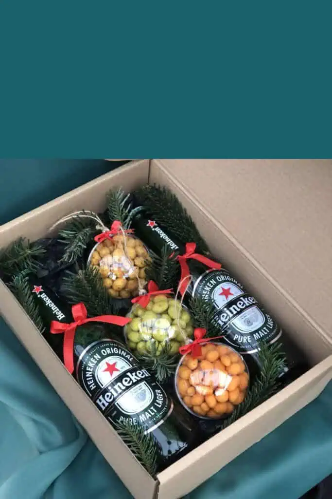 DIY Christmas gift basket idea featuring Heineken beer bottles, clear ornament balls filled with snacks, pine sprigs, and red ribbon inside a cardboard box. A creative and affordable holiday gift basket idea for men.