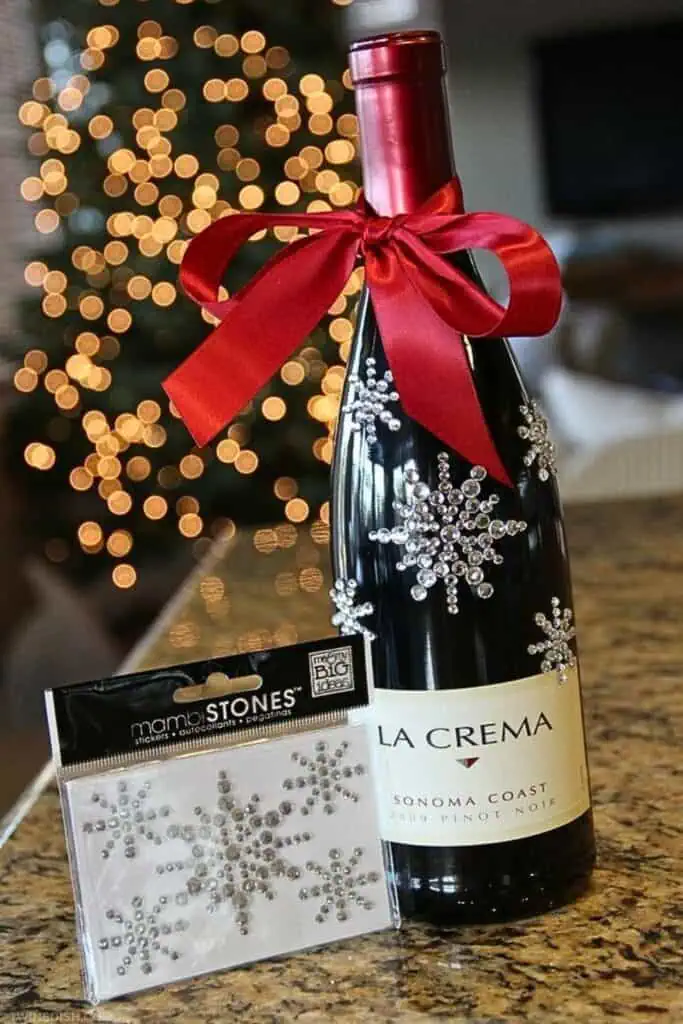 DIY Christmas wine bottle gift decorated with rhinestone snowflake stickers and a red satin bow. A glam and budget-friendly holiday gift or party hostess idea that’s trending for Christmas this year.