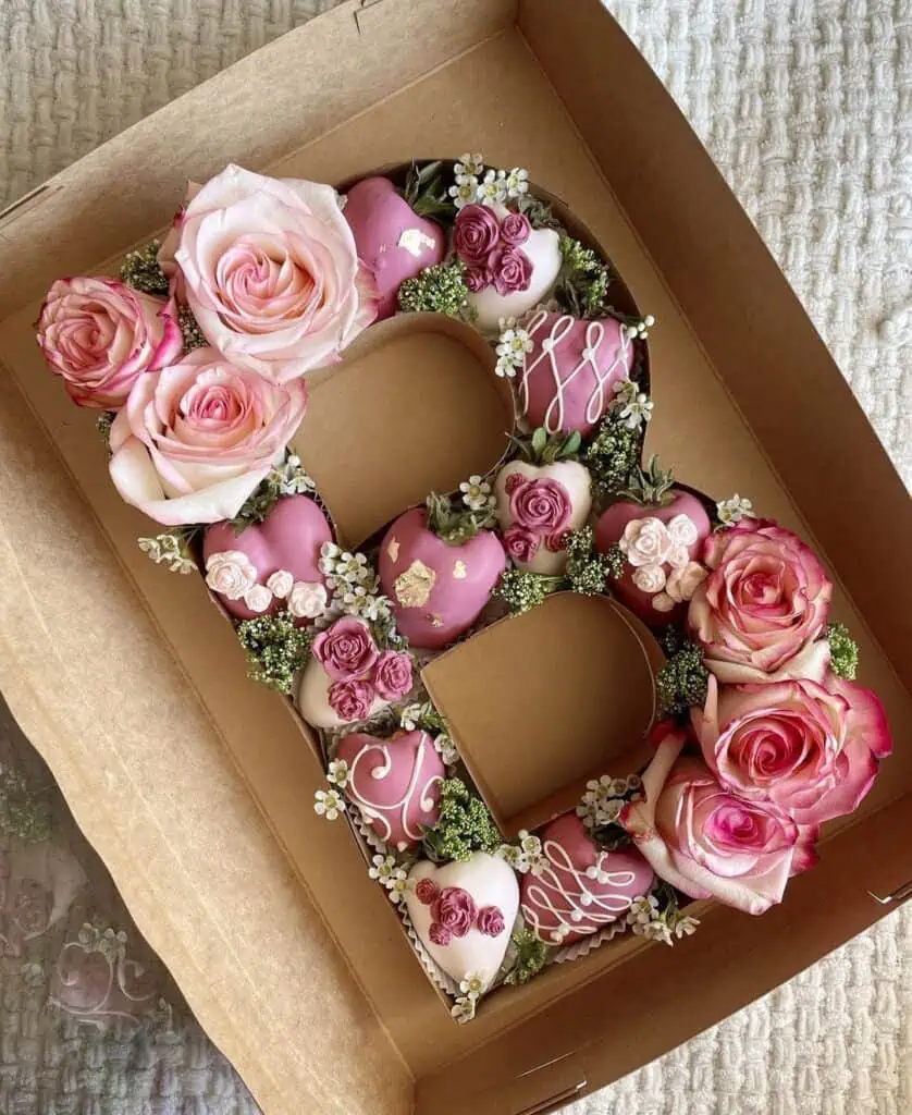 Letter-shaped Valentine’s Day gift box filled with pink roses, chocolate-covered strawberries, and decorative sweets arranged in a brown kraft box.