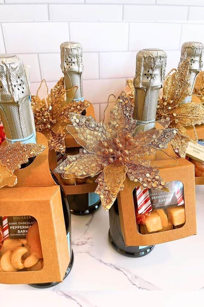Champagne bottles decorated for Christmas with large gold poinsettia flowers and attached gift boxes containing chocolate peppermint bark and assorted snacks.