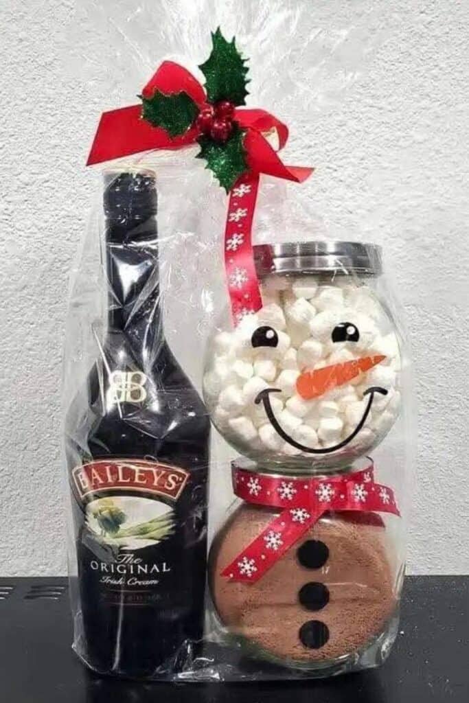 DIY Christmas gift basket idea featuring a Baileys Irish Cream bottle wrapped with a snowman hot cocoa kit. The snowman is made from jars filled with cocoa mix and marshmallows, decorated with a carrot nose, smile, and red holiday ribbon scarf, perfect for gifting.