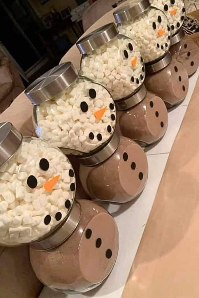 A row of DIY snowman gift jars made from Dollar Store containers, with the bottom jar filled with hot cocoa mix and the top filled with mini marshmallows. Black paper cutouts form the eyes, mouth, and buttons, and an orange paper triangle creates the nose to resemble a snowman. These easy and inexpensive hot chocolate snowmen make adorable Christmas gifts for teachers, coworkers, neighbors, or gift basket fillers.