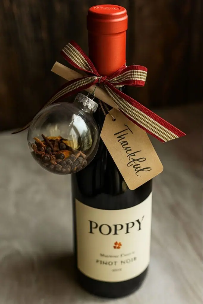 Wine bottle decorated with a plaid ribbon bow, “Thankful” gift tag, and a clear ornament filled with dried spices for mulling.