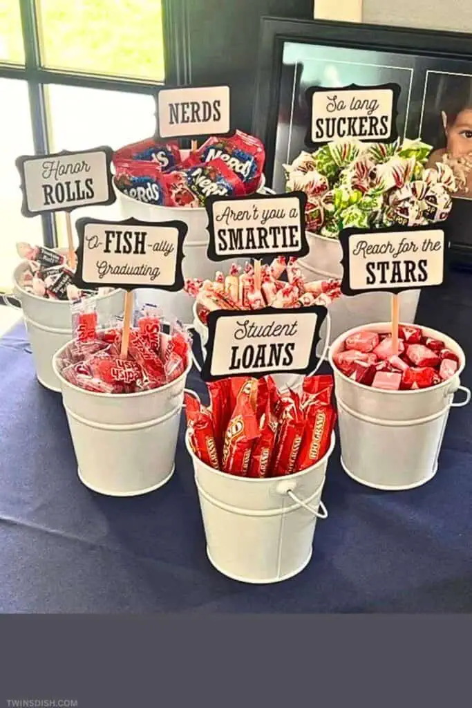 DIY graduation party candy bar display featuring white buckets filled with themed candy and hilarious pun signs like “Honor Rolls” and “Student Loans.” A creative and budget-friendly food table idea trending for 2025 grad celebrations.