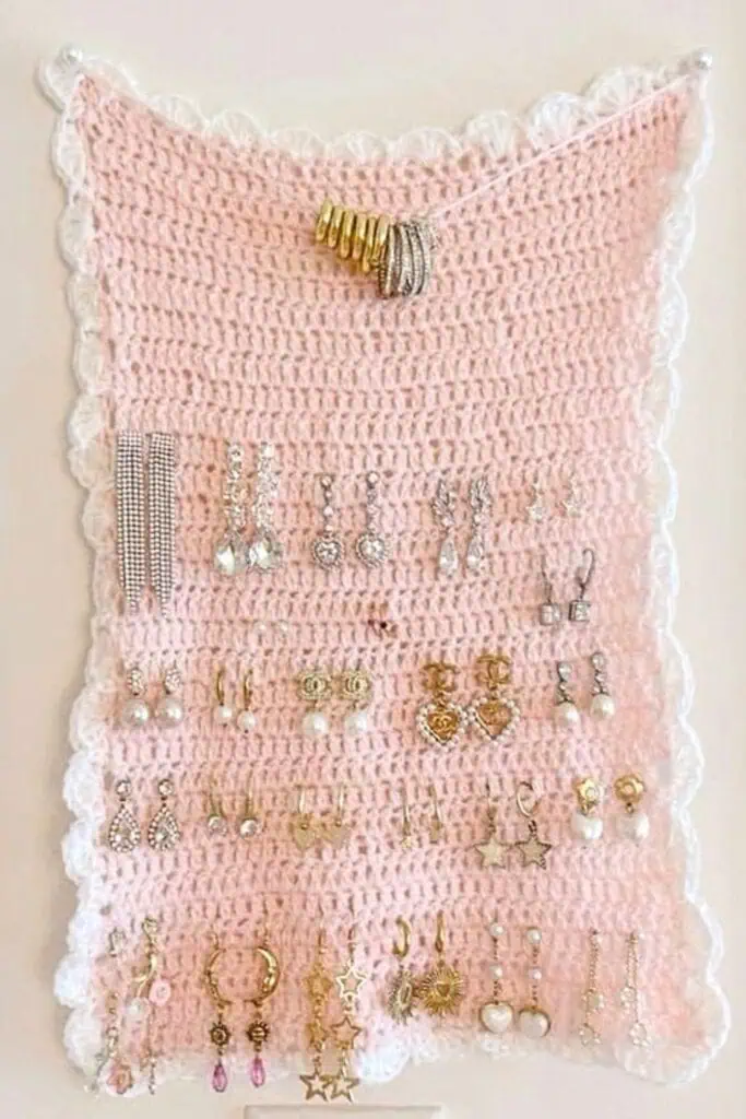 Pink and white crocheted jewelry holder hanging on a wall, with multiple rows of earrings neatly displayed. Handmade using a simple double crochet stitch and scalloped edge border.