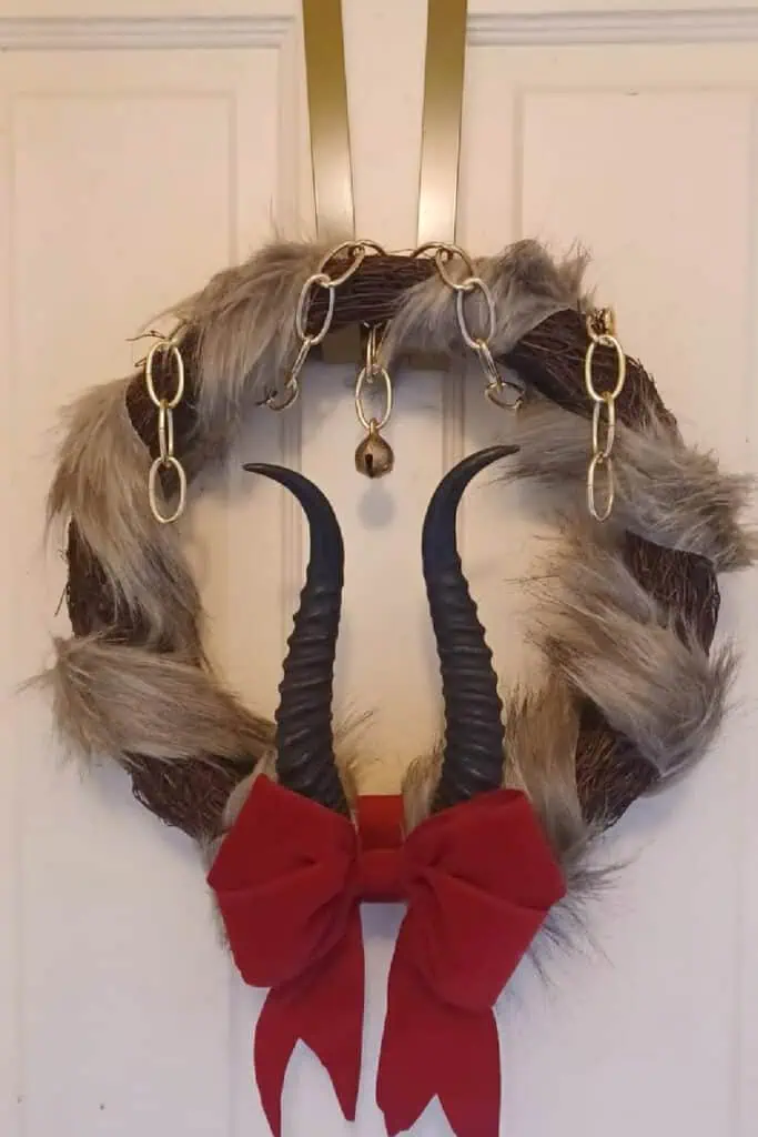 A DIY Krampus Christmas wreath featuring black horns, faux fur, a red bow, gold chain, and bell hanging on a WHITE wooden door.