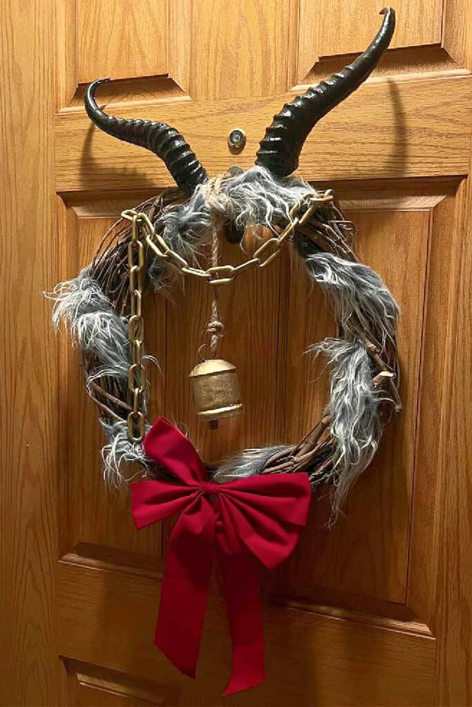 A GOTHIC DIY Krampus Christmas wreath featuring black horns, faux fur, a red bow, gold chain, and bell hanging on a wooden door.