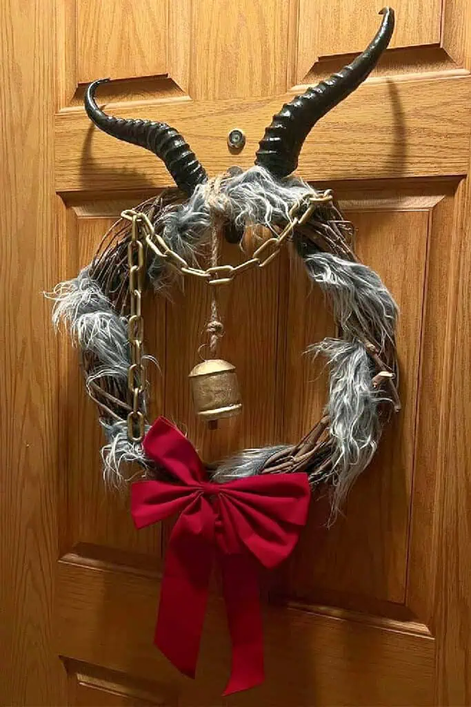 A GOTHIC DIY Krampus Christmas wreath featuring black horns, faux fur, a red bow, gold chain, and bell hanging on a wooden door.