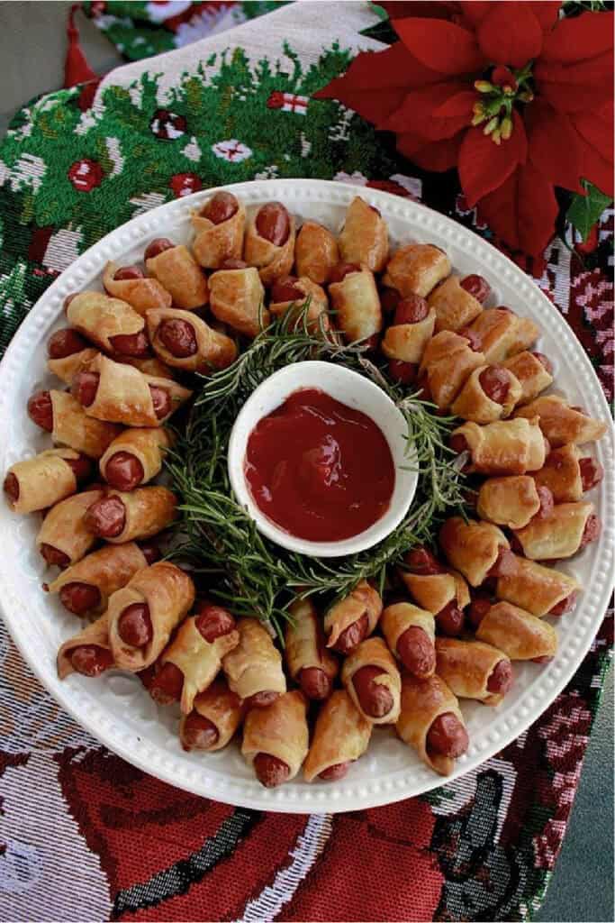 Pigs in a blanket wreath made with mini sausages wrapped in crescent dough, baked until golden and arranged in a festive Christmas wreath with rosemary and dipping sauce.