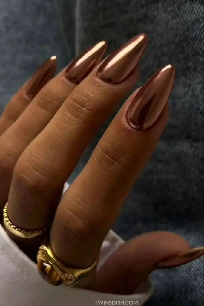 Fall 2025 nail trend featuring almond-shaped copper chrome nails with gold rings and soft denim background