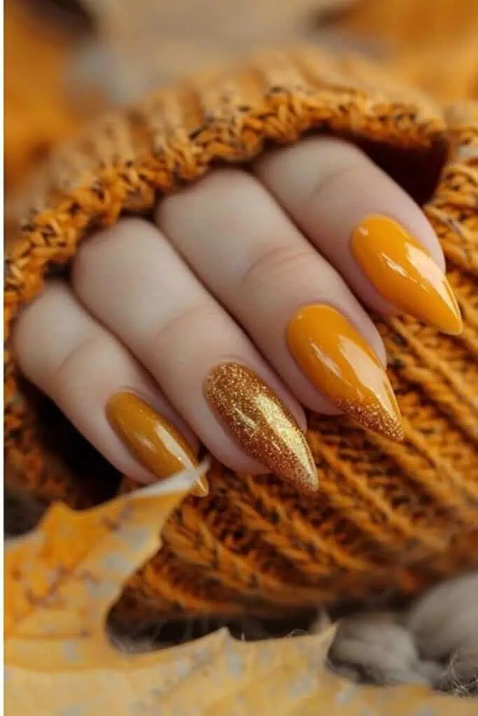 Close-up of trendy almond-shaped fall nails in a glossy burnt orange and golden glitter ombré design, styled with cozy knitwear and autumn leaves. A perfect example of the most pinned nail ideas for fall 2025