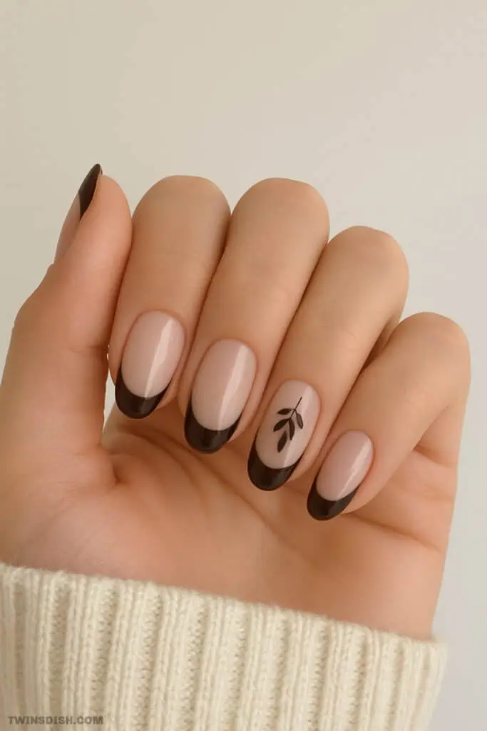 Close-up of a woman's hand with almond-shaped nude nails featuring dark brown French tips and a delicate black leaf design on the ring finger, styled for a modern fall manicure