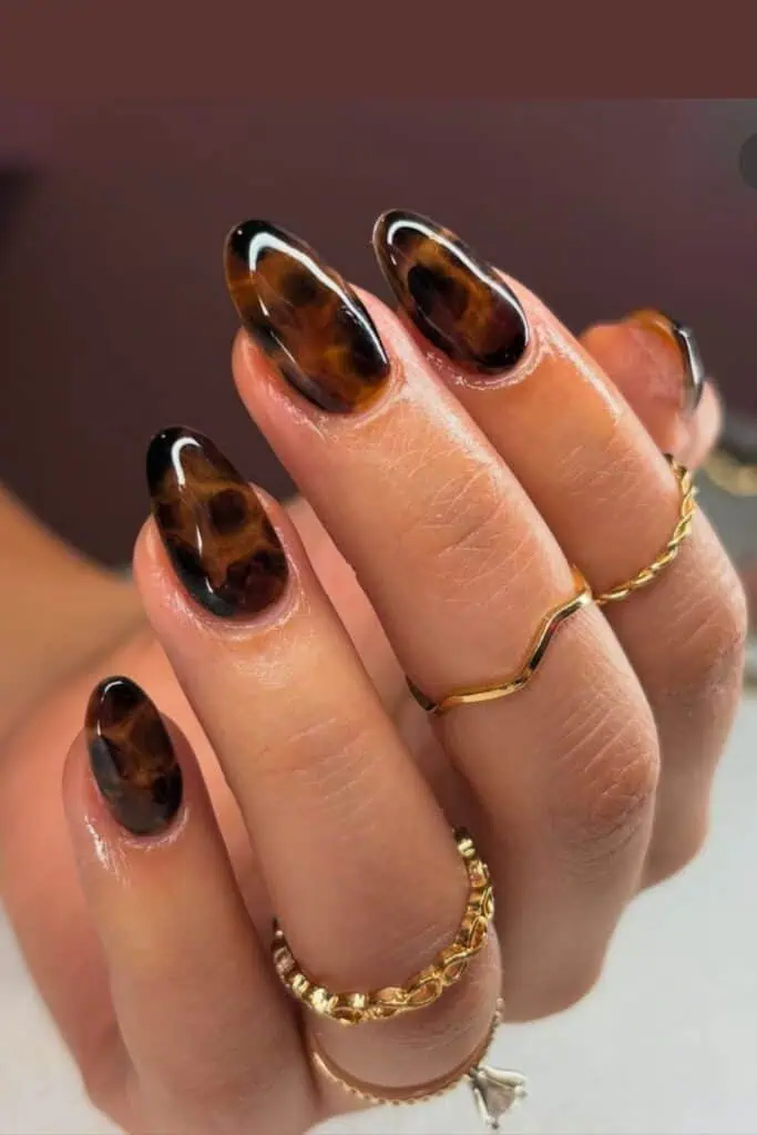 Close-up of glossy oval nails in a tortoise shell design with brown and amber tones, styled with gold rings.