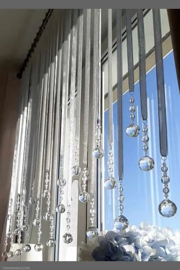 Elegant DIY Christmas window decor with sheer ribbon strands and hanging crystal beads reflecting sunlight. This luxe, glam window styling idea is trending for holiday decorating this year, perfect for creating a magical winter wonderland effect indoors.