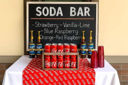Outdoor boys’ graduation party soda bar with Coca-Cola crate, chalkboard sign, red cups, and classic drink syrups – fun and budget-friendly DIY party idea for High School, and College Grad guys.