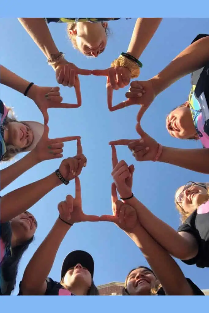 Group of friends forming a crucifix or cross shape with their hands while standing in a circle, photographed from below against a clear blue sky—symbolic and creative photo capturing unity, faith, and friendship.