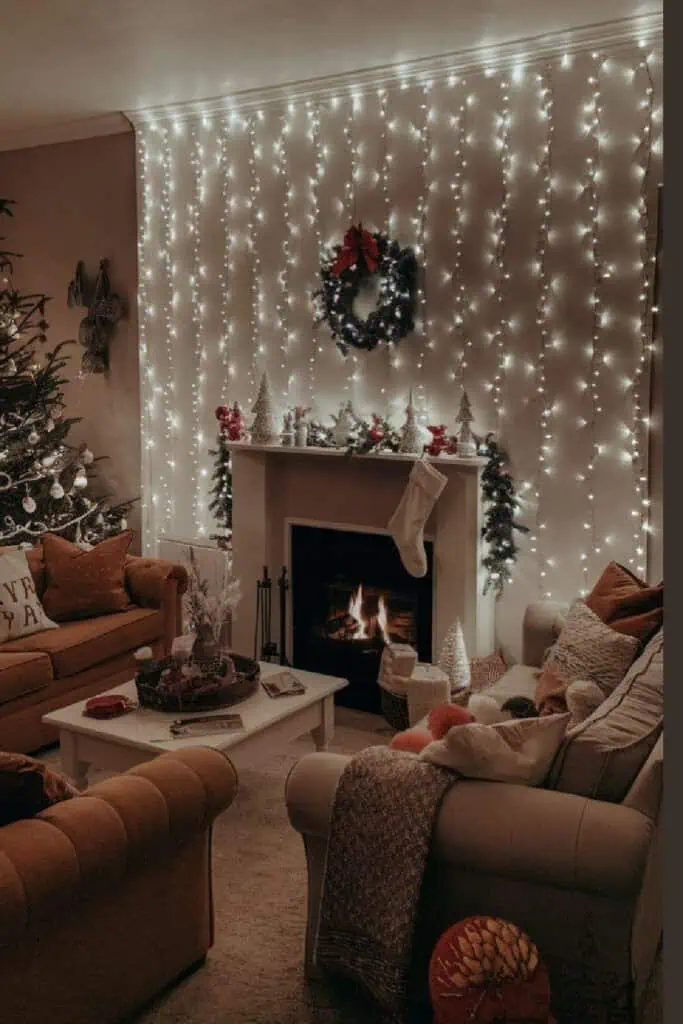 A cozy Christmas living room decorated with warm neutral tones, a lit fireplace, a decorated tree with white ornaments, and a wall of cascading fairy lights behind a white mantle. The mantel is styled with miniature trees, garland, and a wreath with a red bow, creating a soft, festive glow. This inviting holiday scene captures the essence of modern cozy Christmas decor trends.