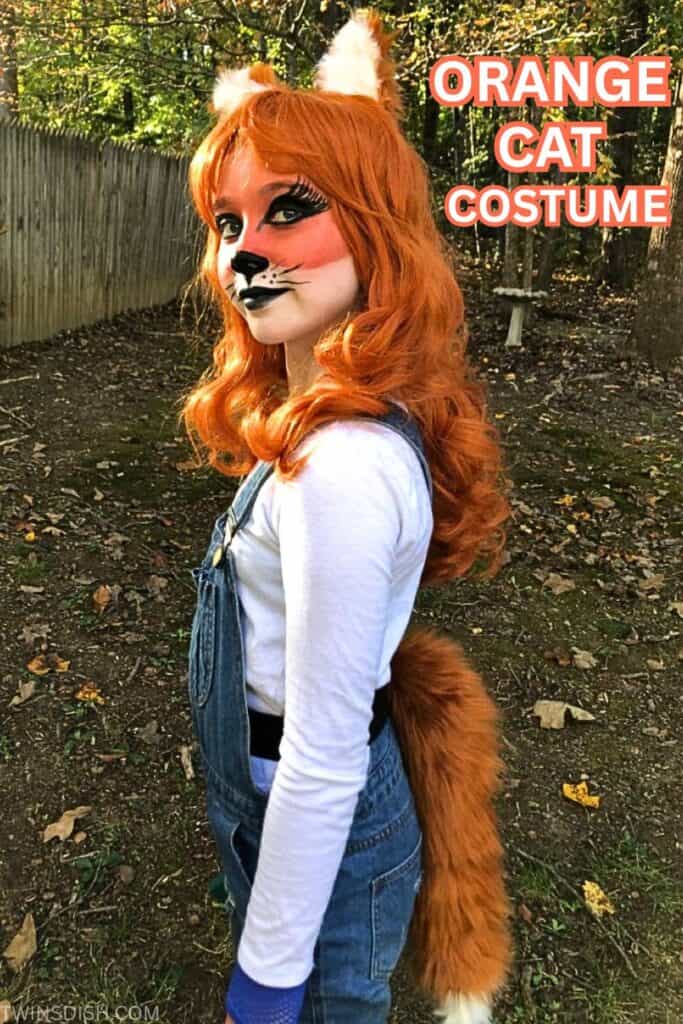 Teen girl wearing a DIY orange cat Halloween costume with dramatic face paint, furry orange ears, long red-orange wig, and a fluffy tail. She’s dressed in denim overalls and a long-sleeve white shirt, standing outside with fall leaves and trees in the background.