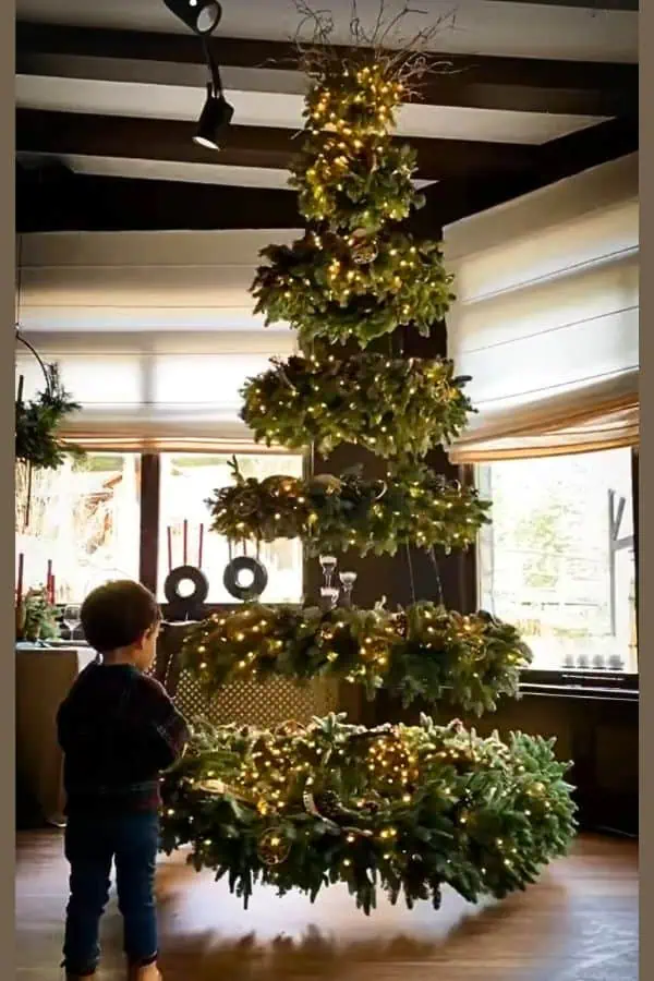 Unique DIY spiral Christmas tree made from floating greenery rings and string lights, creating a magical illusion of a suspended tree in a cozy living room. A little child admires the twinkling holiday decor. Perfect for minimalist or modern Christmas decorating ideas this year