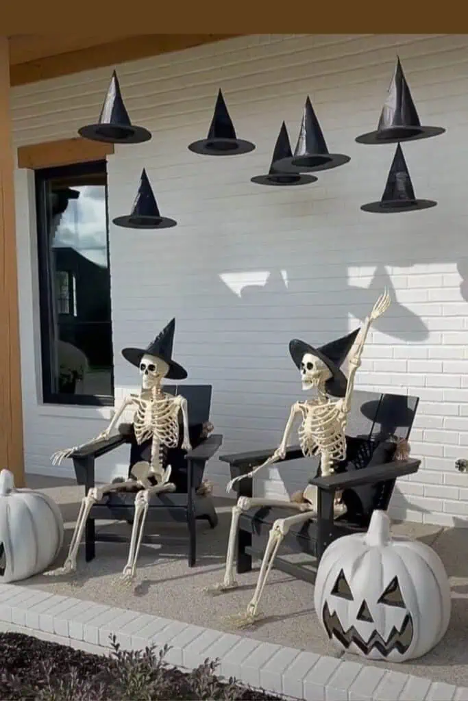 Outdoor Halloween porch decor featuring two skeletons wearing witch hats seated on chairs with floating witch hats above and white pumpkin decorations. A spooky and fun Halloween front porch idea.