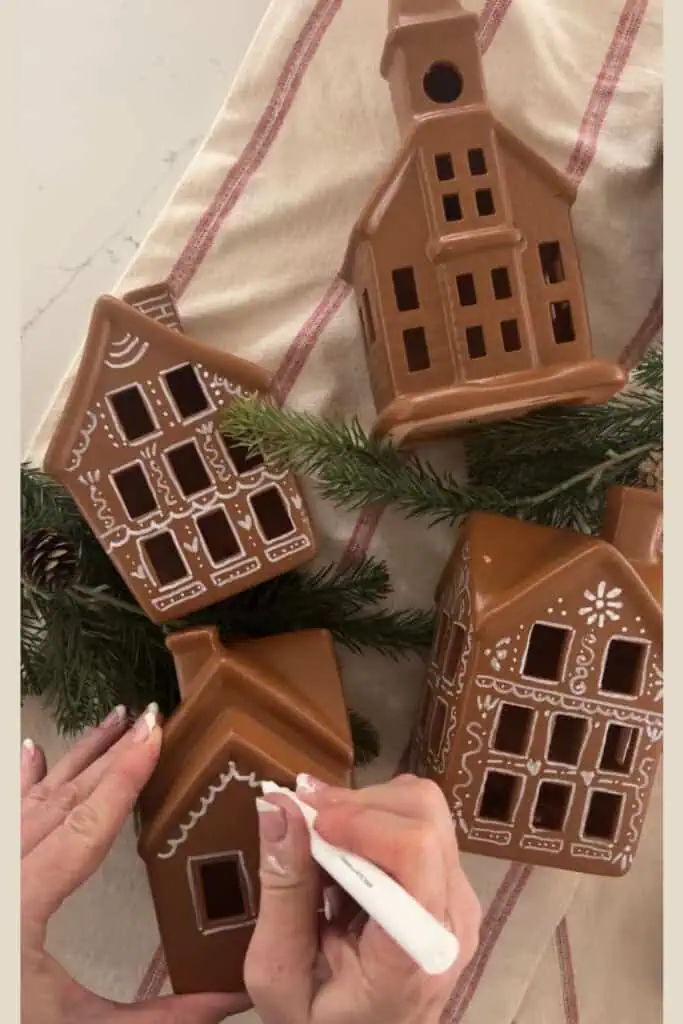 Hands drawing white icing details on brown ceramic village houses to make gingerbread‑style Christmas decor, with pine branches on a striped cloth.
