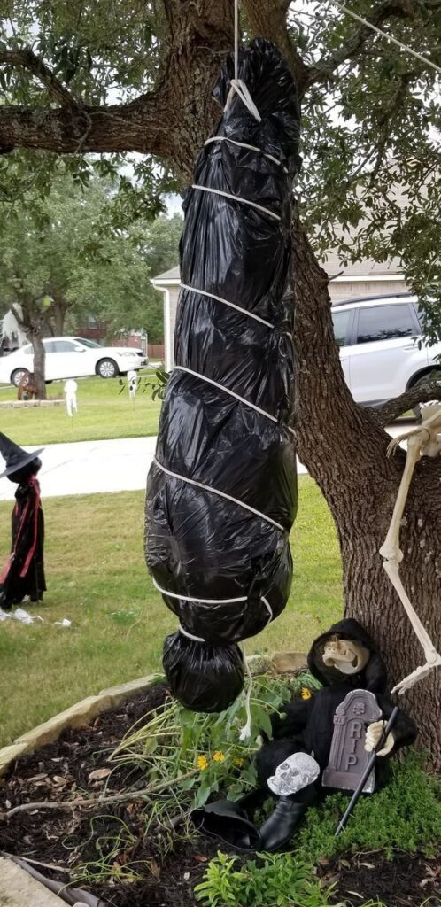 DIY Halloween body bag decoration hanging from a tree outdoors beside skeleton and grim reaper props for spooky yard decor.