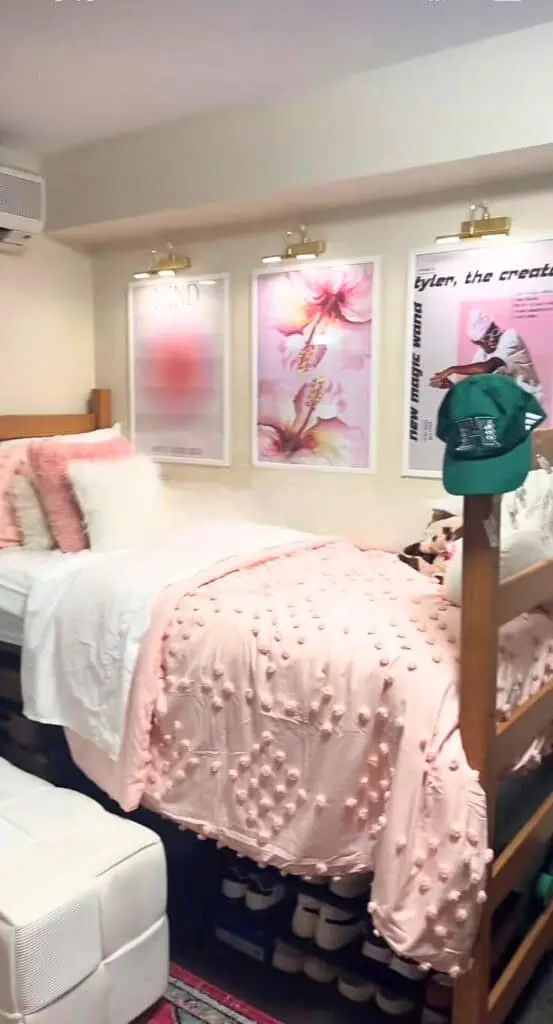 Chic dorm gallery wall with framed prints and gold lighting over pink bedding, styled to look expensive on a budget.