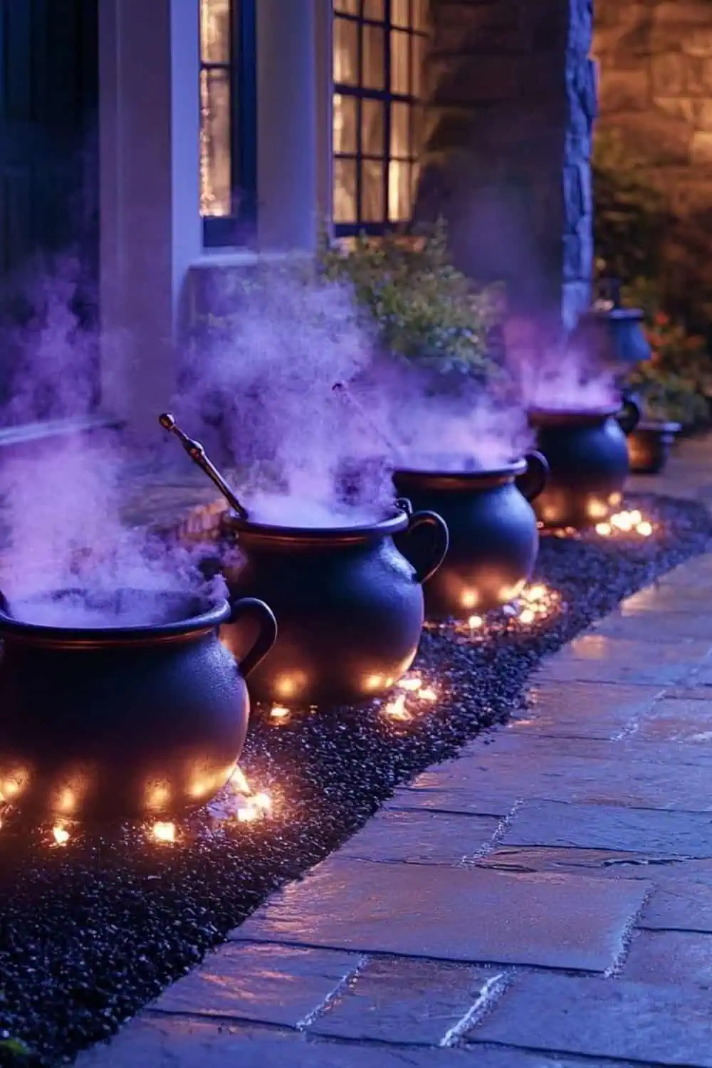 Steaming black witch cauldrons with purple fog line a driveway at night, glowing with tiny pathway lights beside a stone porch; spooky DIY outdoor Halloween decor.