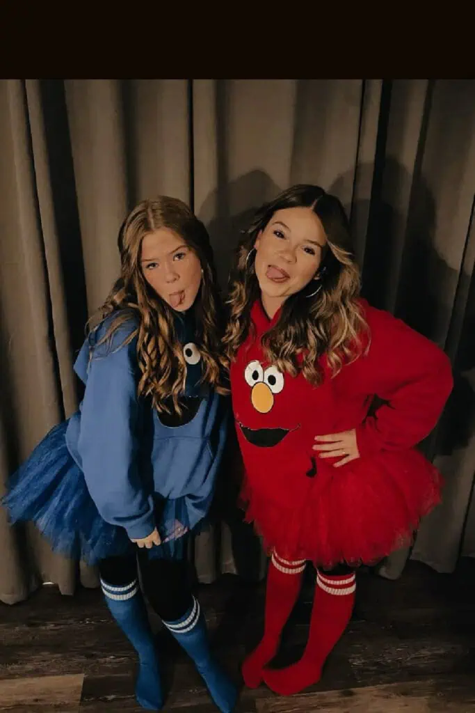 Two girls dressed in duo Halloween costumes as Elmo and Cookie Monster. The Elmo costume features a red hoodie with a tutu and matching socks, while the Cookie Monster costume includes a blue hoodie with a tutu and striped socks. Cute and creative Sesame Street inspired Halloween costume idea for best friends.