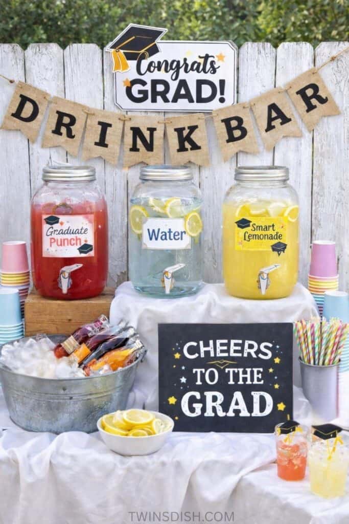 Graduation party drink bar with glass dispensers for punch, water, and lemonade, decorated with a “Cheers to the Grad” sign, cups, and colorful straws.