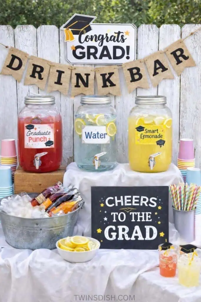 Graduation party drink bar with glass dispensers for punch, water, and lemonade, decorated with a “Cheers to the Grad” sign, cups, and colorful straws.