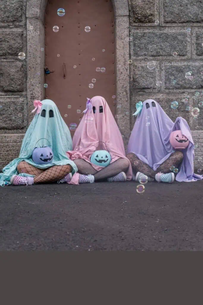 Pastel ghost trio Halloween costumes with pink, blue, and purple sheets, bows, fishnet tights, sneakers, and matching pumpkin buckets. Cute and creative group costume idea.
