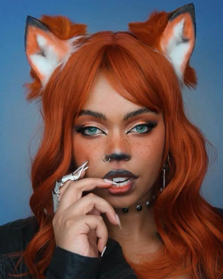 Close-up of a woman wearing an orange cat Halloween makeup look with dramatic eyeliner, a dark contoured lip, orange faux freckles, and a blackened feline nose. She wears orange wavy hair, realistic cat ears, and statement rings with a dark choker necklace, creating a bold and glamorous costume.
