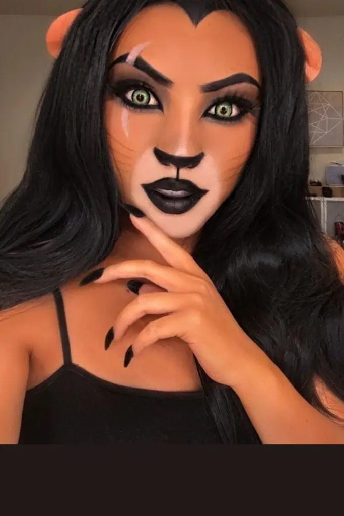 Halloween makeup inspired by Scar from The Lion King with orange and black face paint, scar detail across the eye, green contact lenses, black lipstick, and lion ears.
