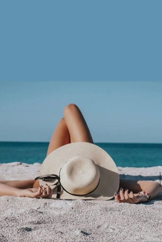 Woman lying on the beach with a straw sunhat covering her face, legs relaxed and propped up toward the ocean—effortlessly chic summer photo with a serene seaside backdrop.