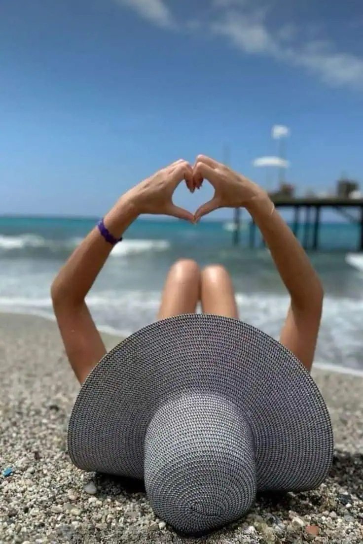 Picture of a woman lying on a pebbled beach facing the ocean, wearing a wide-brimmed sun hat and forming a heart shape with their hands against a blue sky backdrop. Waves roll gently onto the shore, and a pier extends into the water in the background.