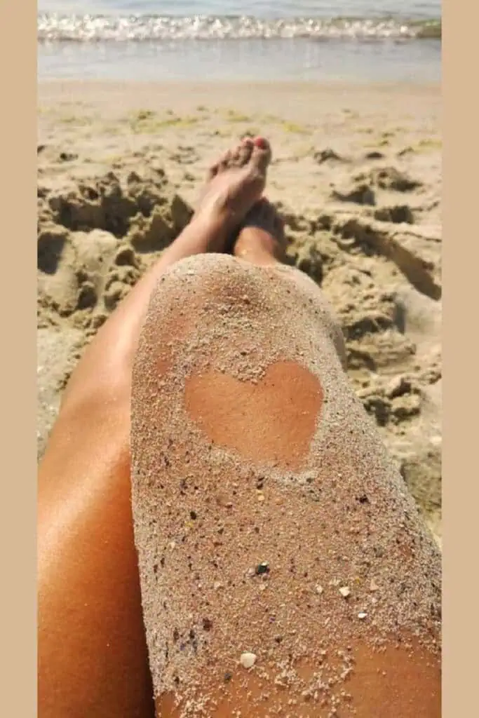 Legs covered in sand with a heart shape showing skin underneath, captured at the beach. Aesthetic, flattering summer photo pose perfect for Instagram or Pinterest.